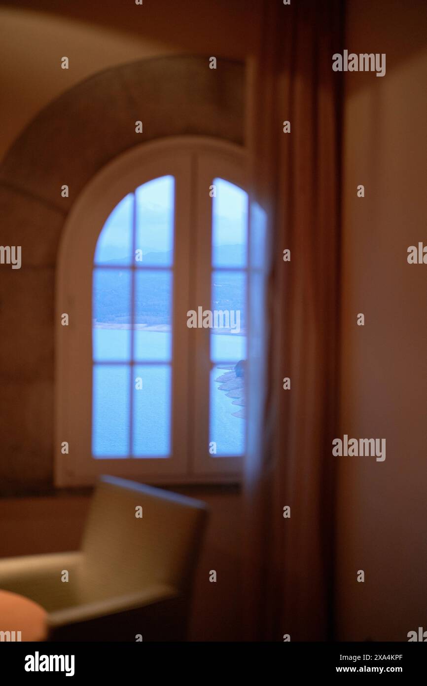 Una finestra ad arco che proietta luce naturale soffusa in una sala con vista parziale di una sedia e di una tenda, creando un'atmosfera serena e tranquilla. Foto Stock