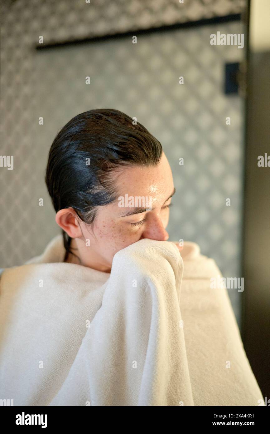 Una giovane donna è vista con i capelli bagnati spazzati indietro, coperti da un asciugamano beige, e davanti a uno sfondo testurizzato grigio scuro. Foto Stock