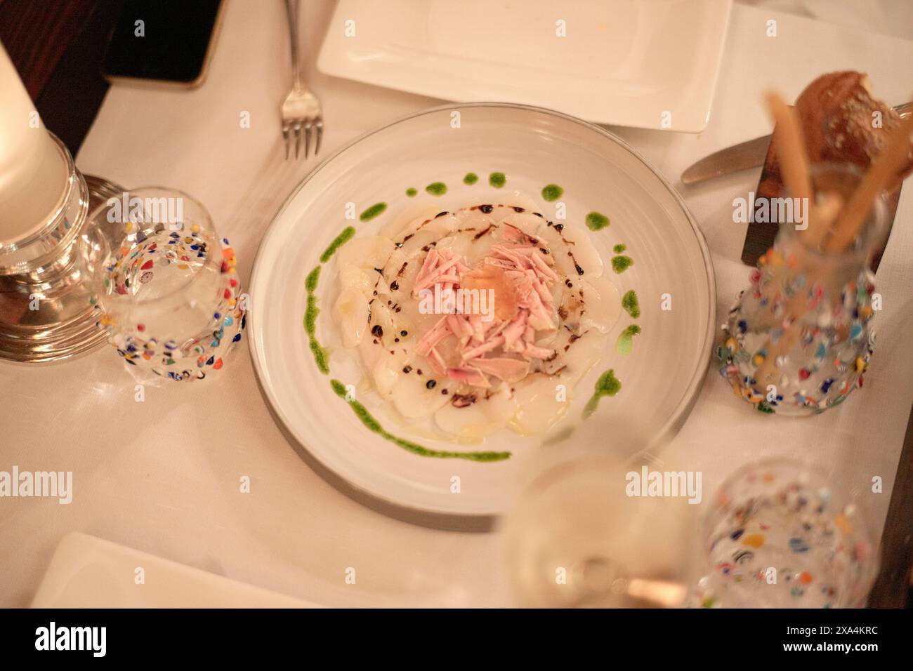 Si tratta di una vista dall'alto di un piatto finemente placcato, possibilmente un'insalata, circondato da eleganti bicchieri su una tovaglia bianca. Foto Stock