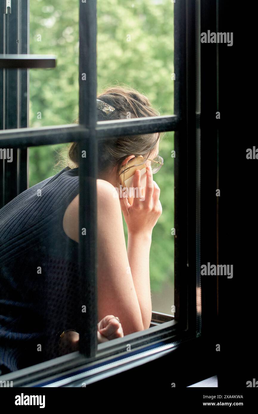 Una persona appoggia la testa sulla mano e guarda fuori da una finestra aperta, con vegetazione visibile sullo sfondo. Foto Stock