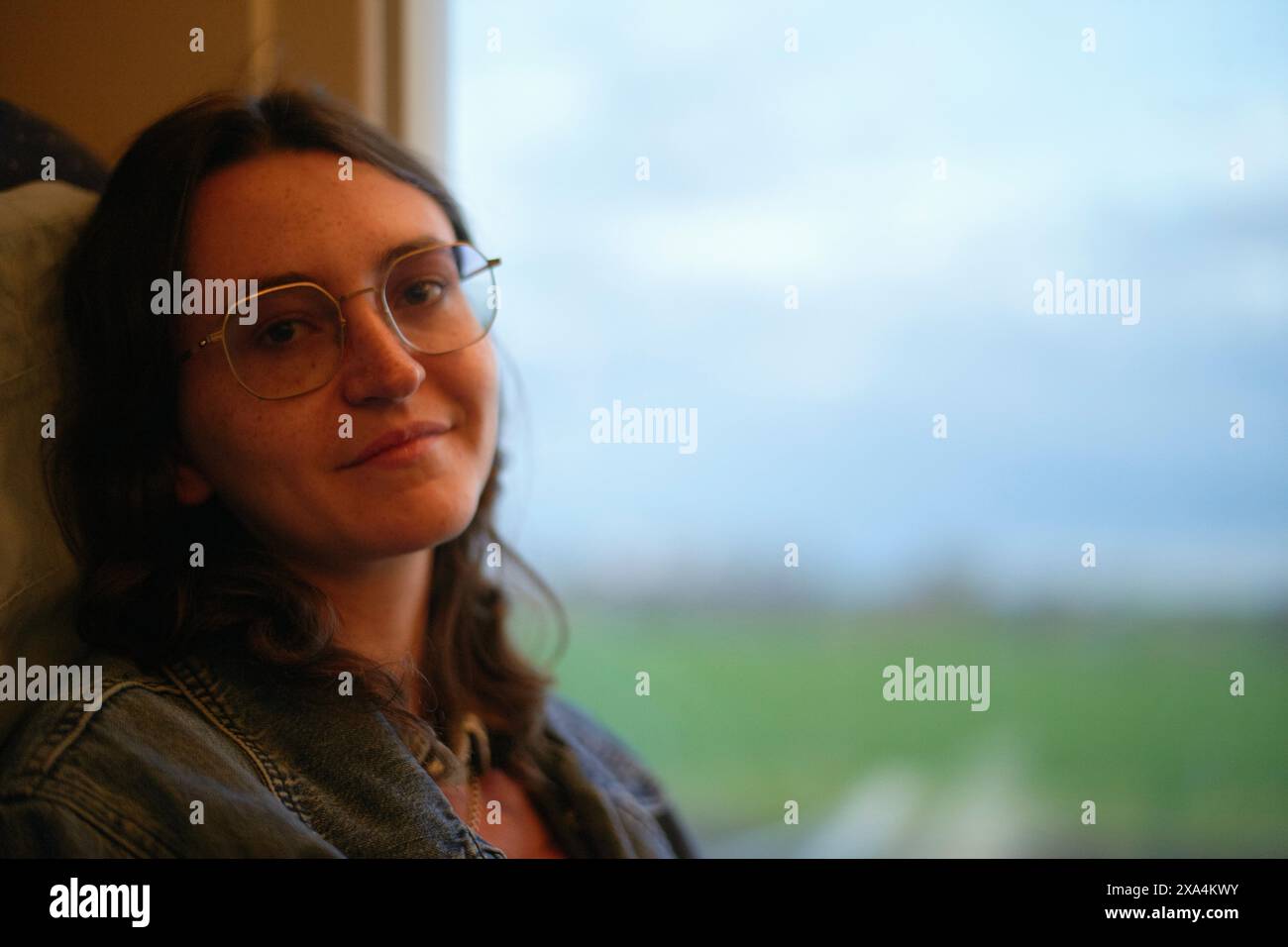 Una donna con gli occhiali è seduta all'interno da una finestra con una vista su un paesaggio sfocato all'esterno, dando un sottile sorriso. Foto Stock