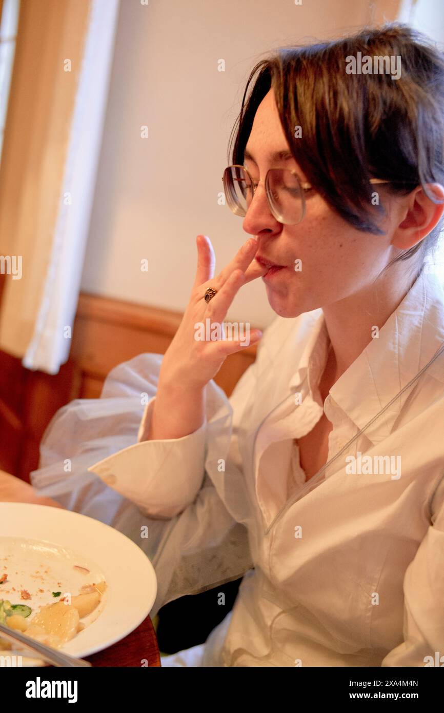 Una persona si siede a un tavolo da pranzo, gustando un pasto, con un piatto che contiene alcuni resti di cibo. Sono vestiti con una camicia bianca e indossano occhiali rotondi, gestando con la mano vicino alla bocca. Foto Stock