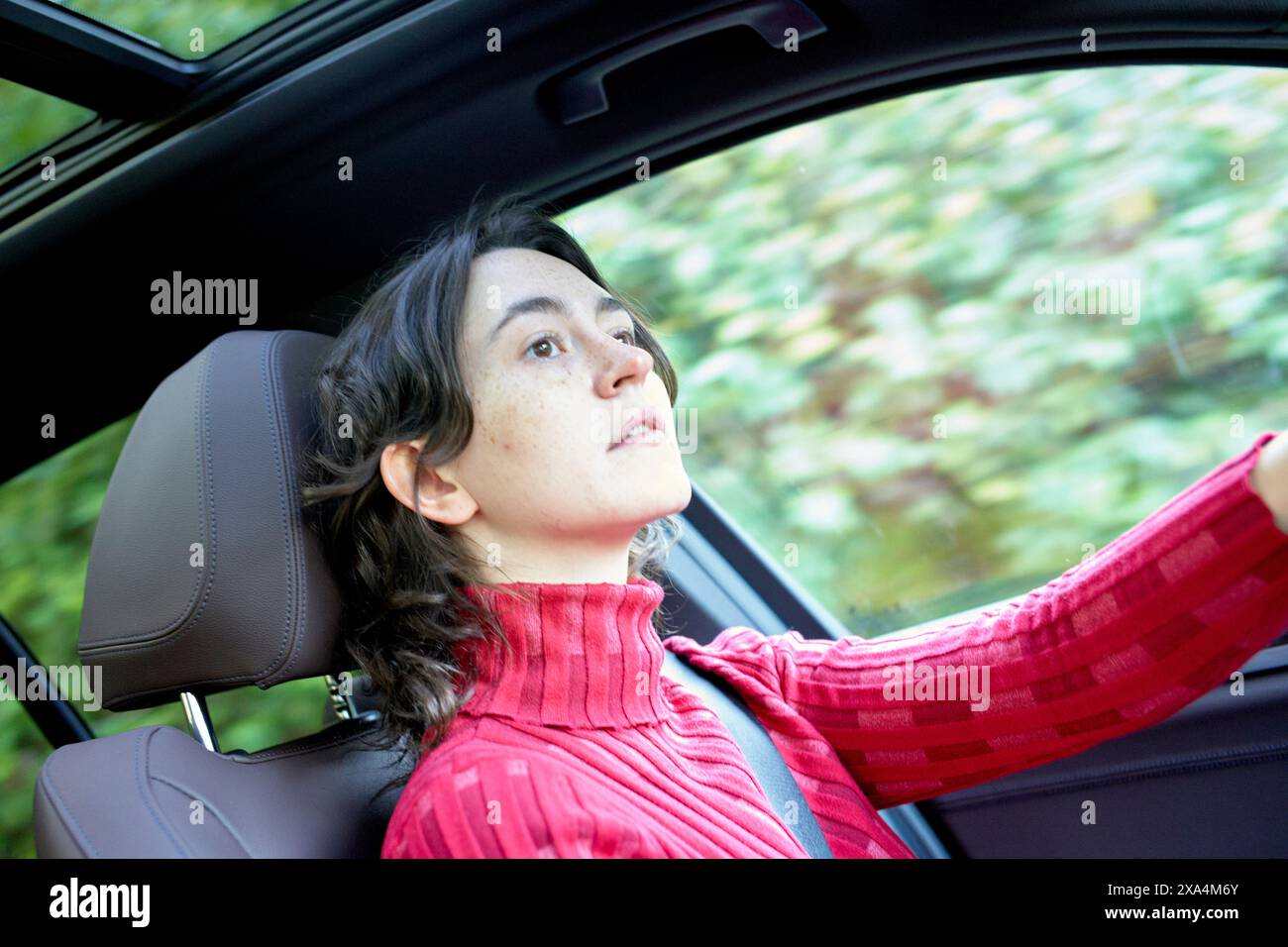 Una donna con il tetto rosso sta guidando un'auto, concentrandosi intensamente sulla strada davanti con alberi visibili sullo sfondo. Foto Stock