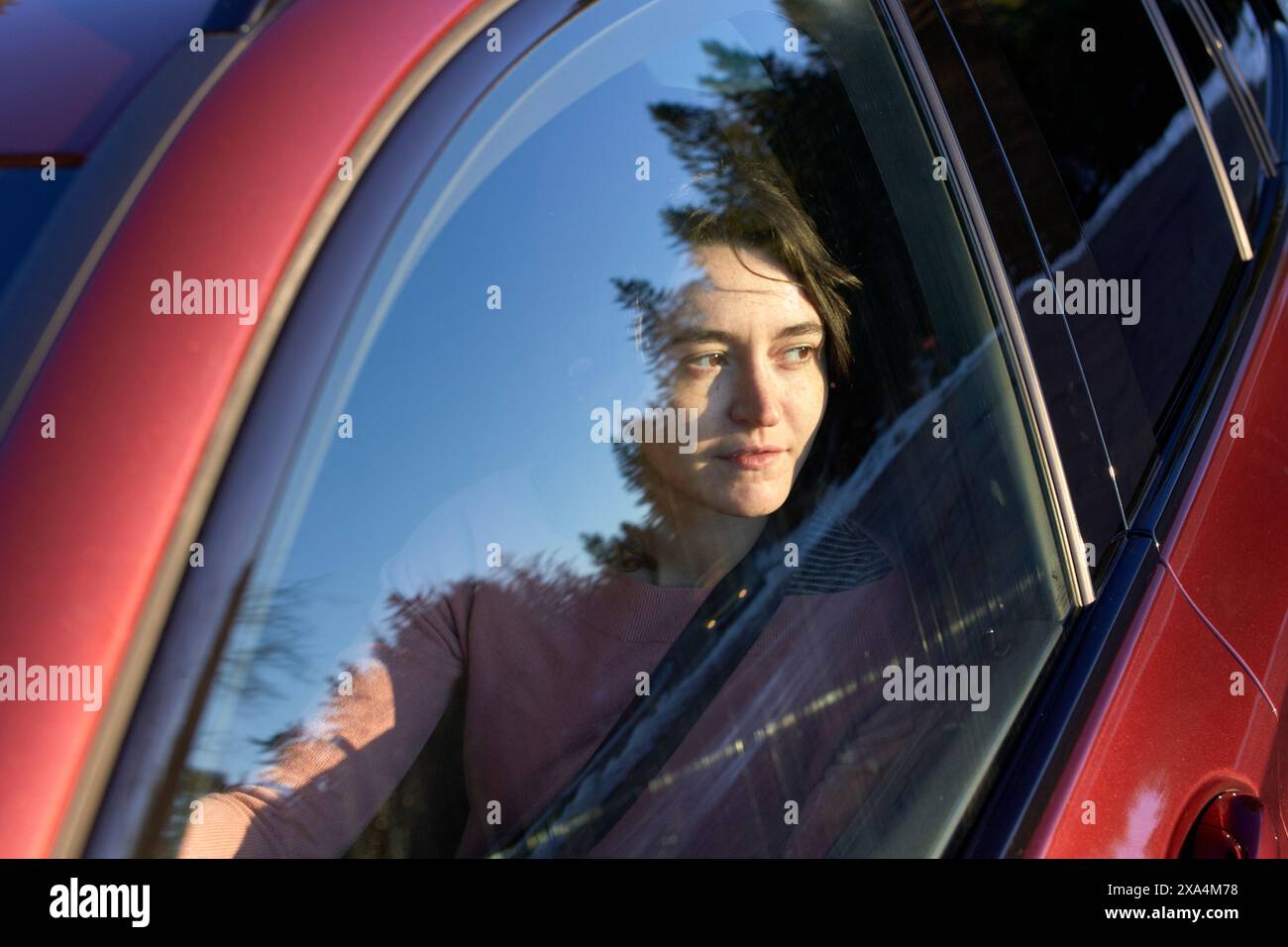 Una persona viene vista attraverso la finestra di un'auto rossa, il suo riflesso si fonde con gli alberi sullo sfondo. Foto Stock