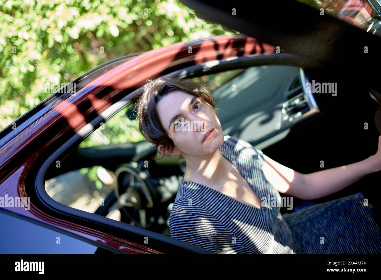 Una persona sta aprendo la portiera dell'auto, guardando verso l'esterno con un'espressione mirata, strisciando leggermente alla luce del sole. Foto Stock