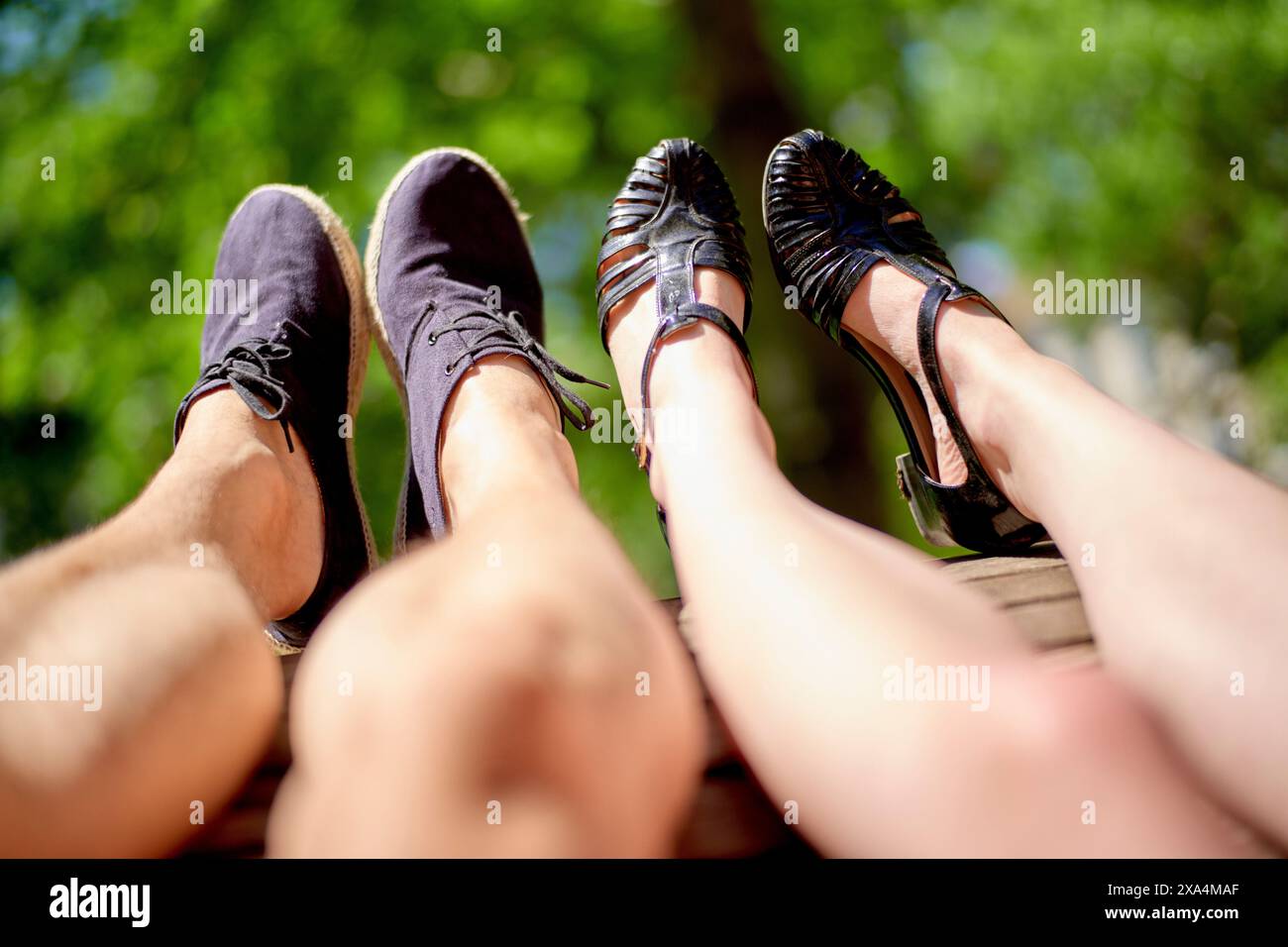 Una coppia ha allungato le gambe verso l'alto su uno sfondo di verde vegetale, mostrando i loro diversi stili di calzature sotto la luce del sole. Foto Stock