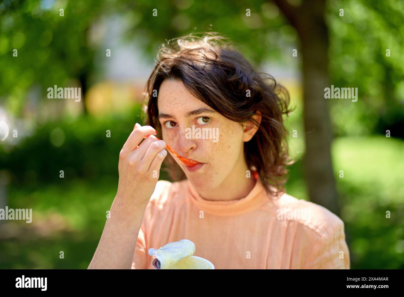 Una persona con lentiggini, che indossa una parte superiore color pesca, sta mangiando una fetta di arancia, con uno sfondo verde sfocato. Foto Stock