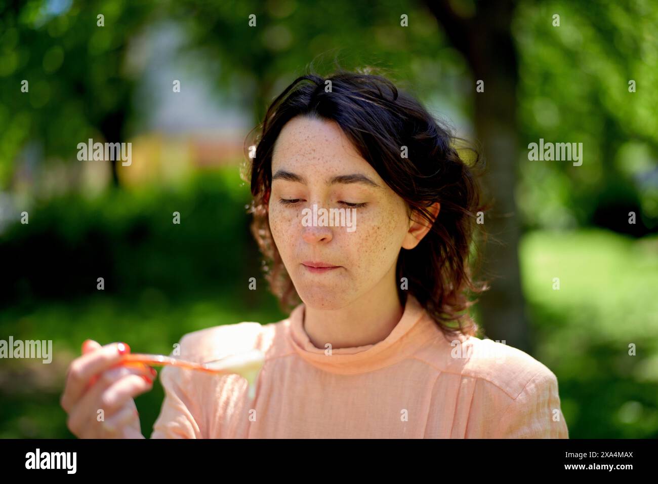 Una giovane donna è all'aperto in una giornata di sole, gli occhi chiusi e sta per mangiare qualcosa da un cucchiaio con vegetazione sfocata sullo sfondo. Foto Stock