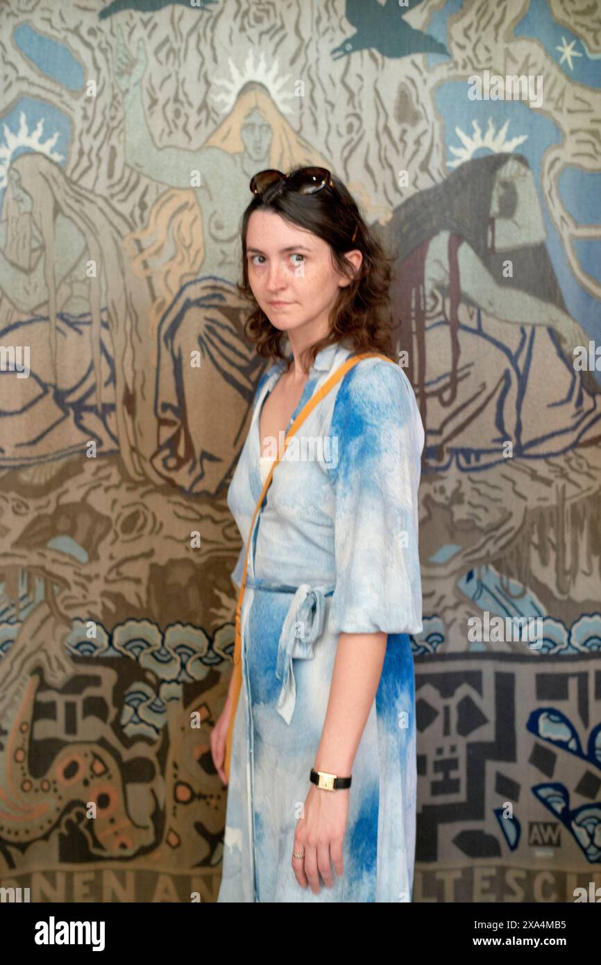 Una donna si erge davanti a un tappeto a parete con motivi blu, indossando un abito blu che si fonde con lo sfondo, accessoriato con una borsa a spalla e occhiali da sole. Foto Stock