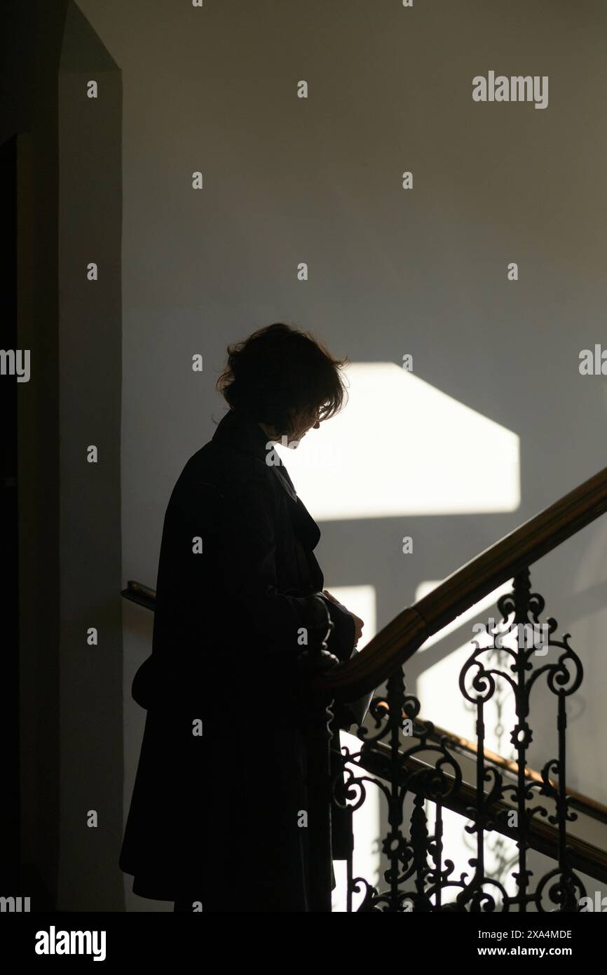 Silhouette di una persona in piedi su una scala con una mano sulla ringhiera, retroilluminata dalla luce brillante di una finestra che proietta ombre. Foto Stock