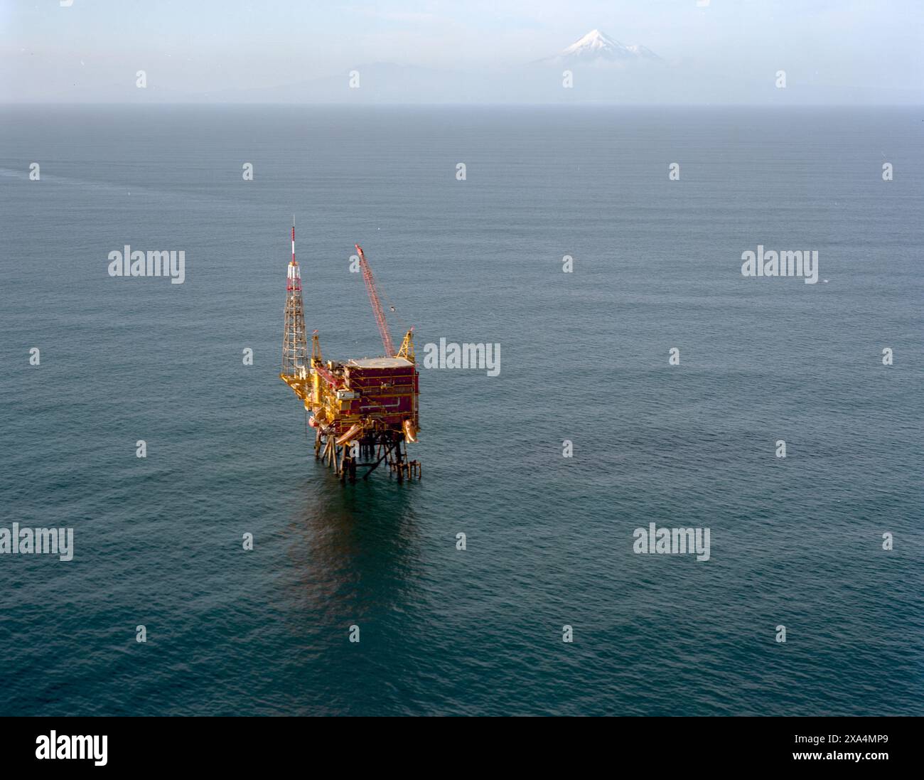 Una piattaforma di perforazione offshore si trova al centro dell'oceano con una montagna distante visibile all'orizzonte sotto un cielo limpido, il Mar di Tasman, nuova Zelanda Foto Stock