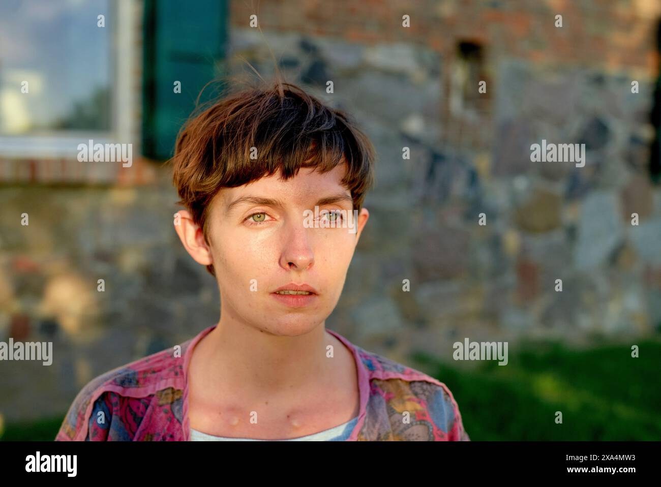 Una donna con un taglio di capelli corto si trova di fronte a un vecchio edificio in mattoni, guardando lateralmente con un'espressione premurosa. Foto Stock