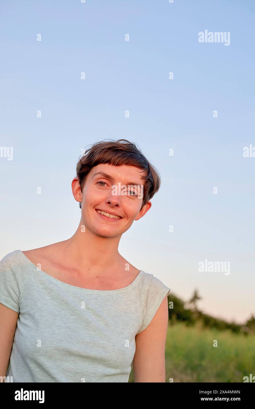 Una donna sorridente con i capelli corti, che indossa una parte superiore di colore chiaro, si erge all'aperto con un cielo limpido sullo sfondo. Foto Stock