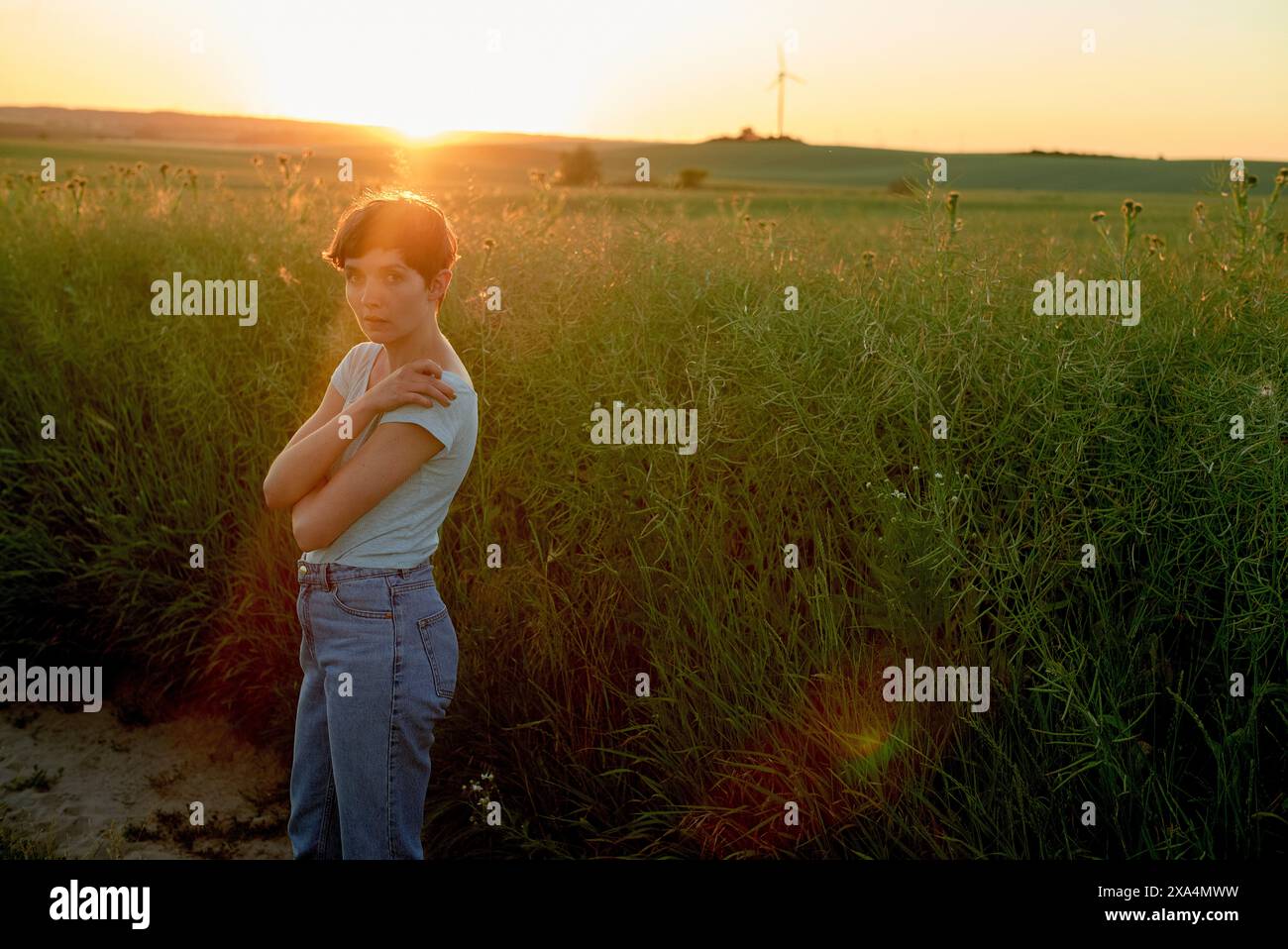 Una giovane donna si trova in un campo verde al tramonto, con il sole che proietta un caldo bagliore e crea uno sfondo sereno con turbine eoliche in lontananza. Foto Stock