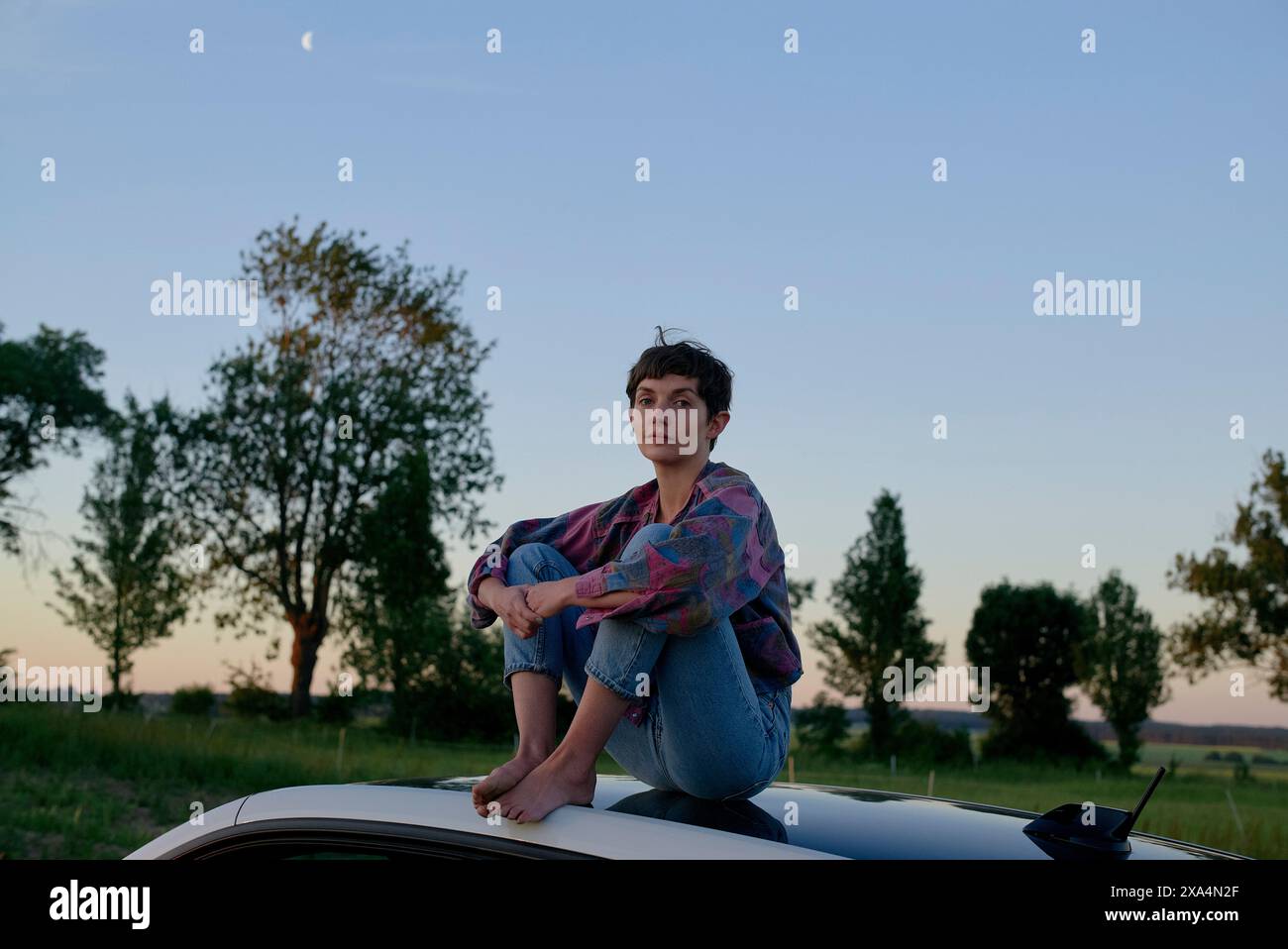 Una giovane donna siede sul tetto di un'auto durante il crepuscolo, con alberi sullo sfondo e una luna crescente visibile nel cielo. Foto Stock