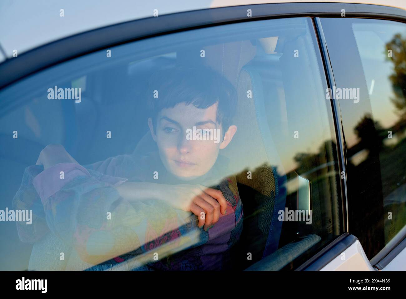 Una giovane donna guarda fuori dal finestrino di un'auto, con un'espressione riflessiva. Il sole tramonta proiettando un caldo bagliore sulla scena. Foto Stock