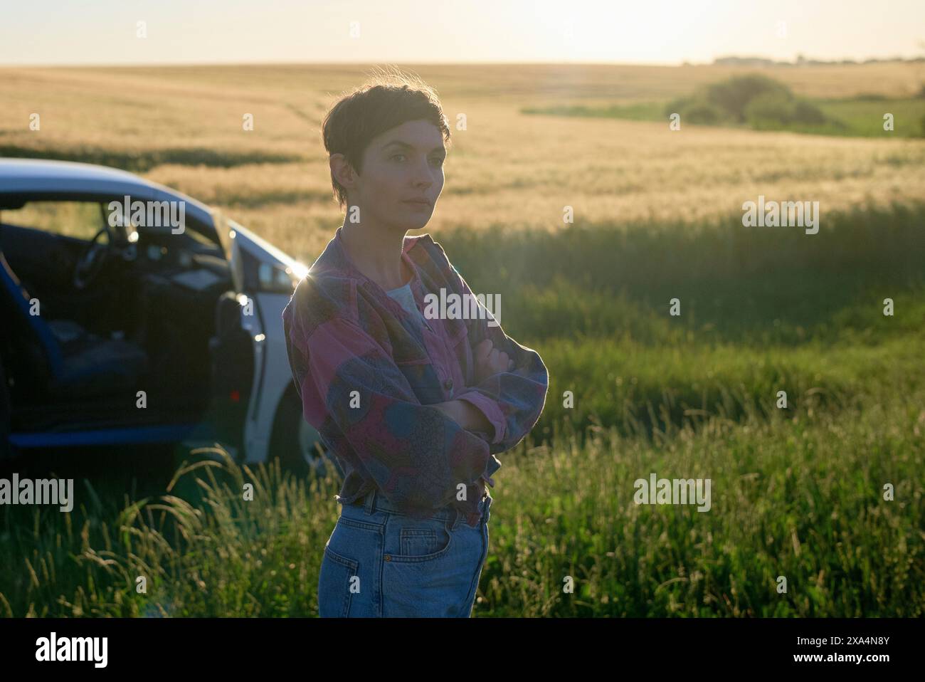 Una giovane donna si trova accanto a un'auto blu in un campo di erba alta dorata, con il sole che tramonta alle spalle e che emette un caldo bagliore. Foto Stock