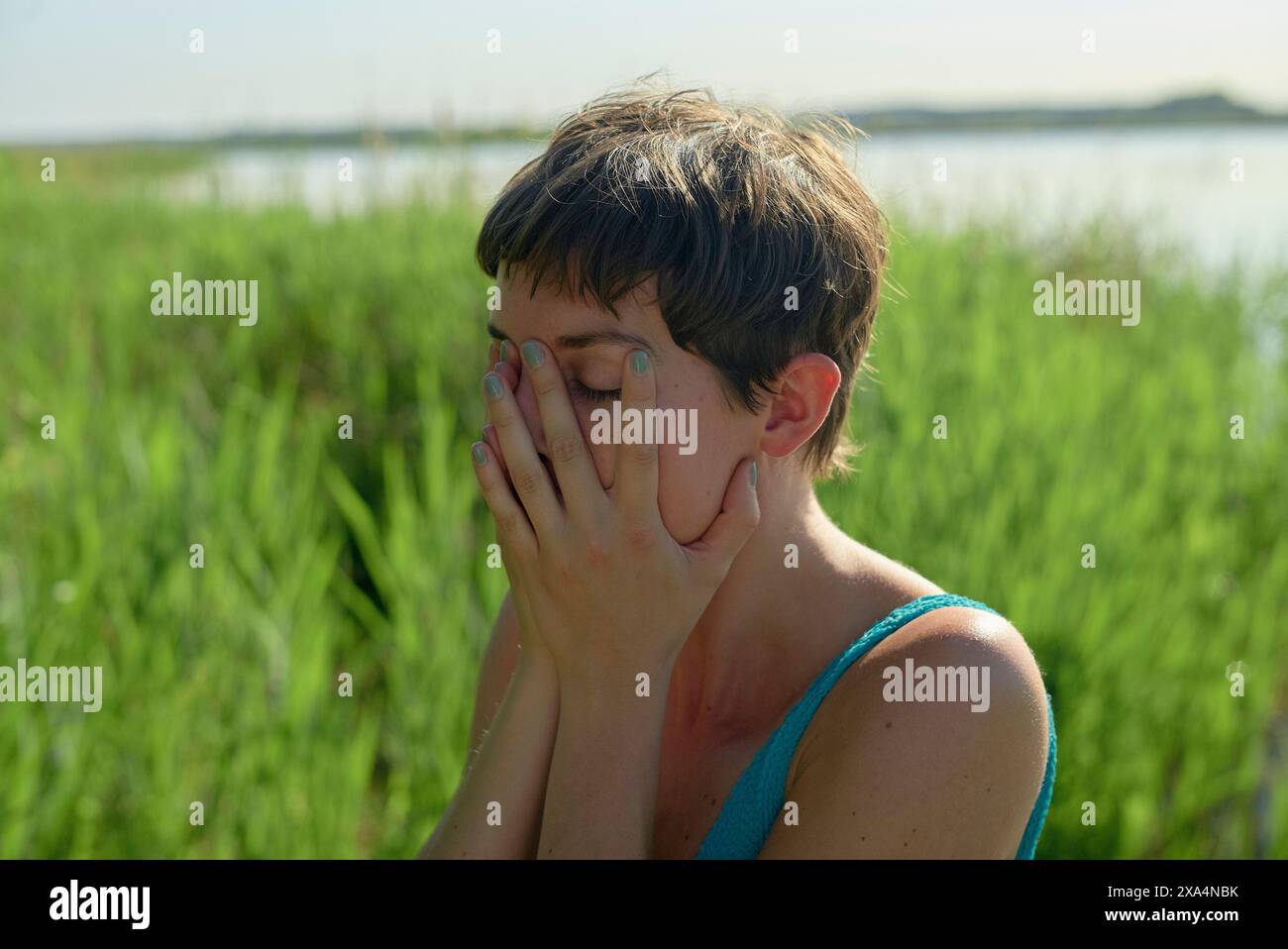 Una giovane donna è seduta all'aperto con gli occhi coperti dalle mani, apparentemente in un momento di angoscia o contemplazione, con un corpo d'acqua e canne verdi sullo sfondo. Foto Stock