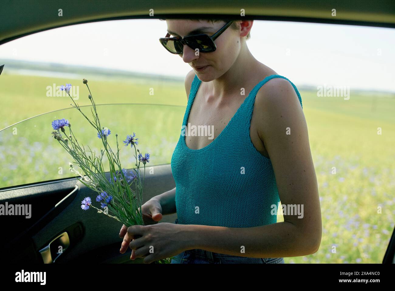 Una donna sta accanto a un'auto, tenendo un mazzo di fiori blu in un campo verde sotto un cielo limpido. Foto Stock