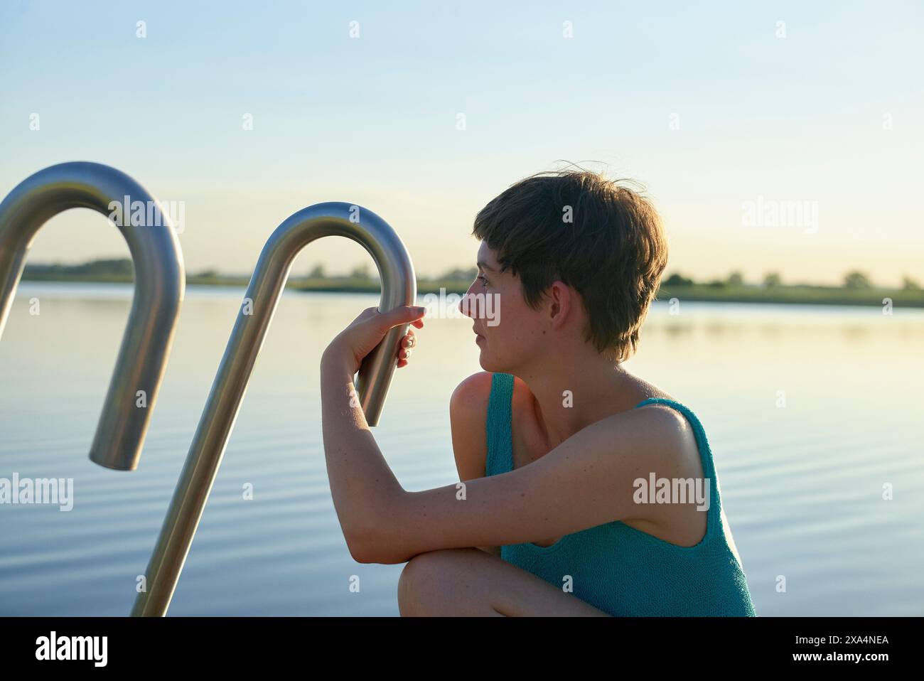 Una giovane donna dai capelli corti sta appoggiando il mento sulla mano mentre è seduta accanto a un tranquillo corpo d'acqua al tramonto, guardando con attenzione in lontananza. Foto Stock
