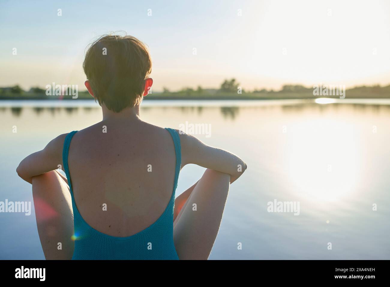 Una giovane donna è seduta accanto a un tranquillo lago al tramonto, con la schiena verso la macchina fotografica e le mani appoggiate sul retro della testa. Foto Stock