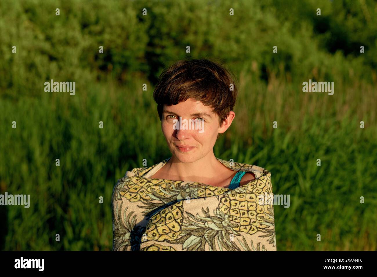 Una donna sorridente con capelli corti in piedi in un campo con alte piante verdi sullo sfondo, durante quella che sembra essere l'ora d'oro del tramonto. Foto Stock