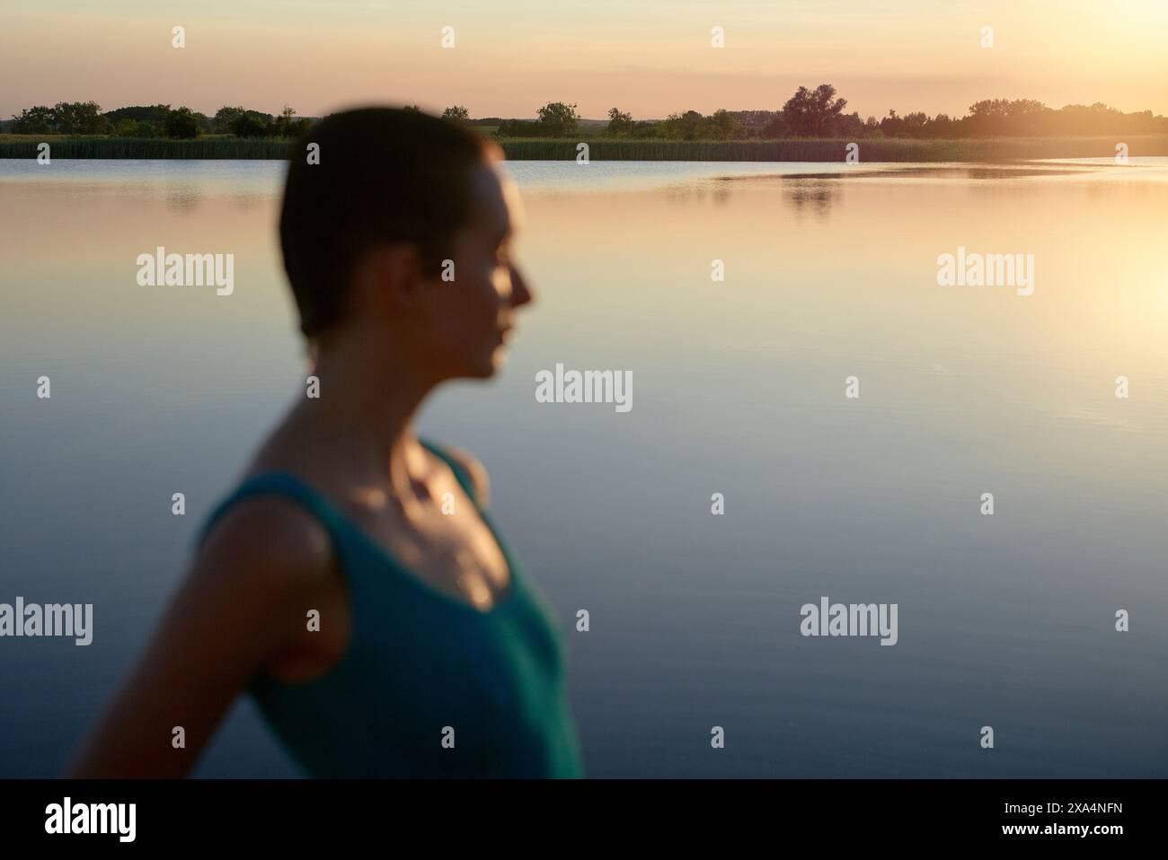 Una persona è in primo piano e gode di una tranquilla vista sul lago durante il tramonto, con un tranquillo riflesso d'acqua e un morbido cielo dorato sullo sfondo. Foto Stock