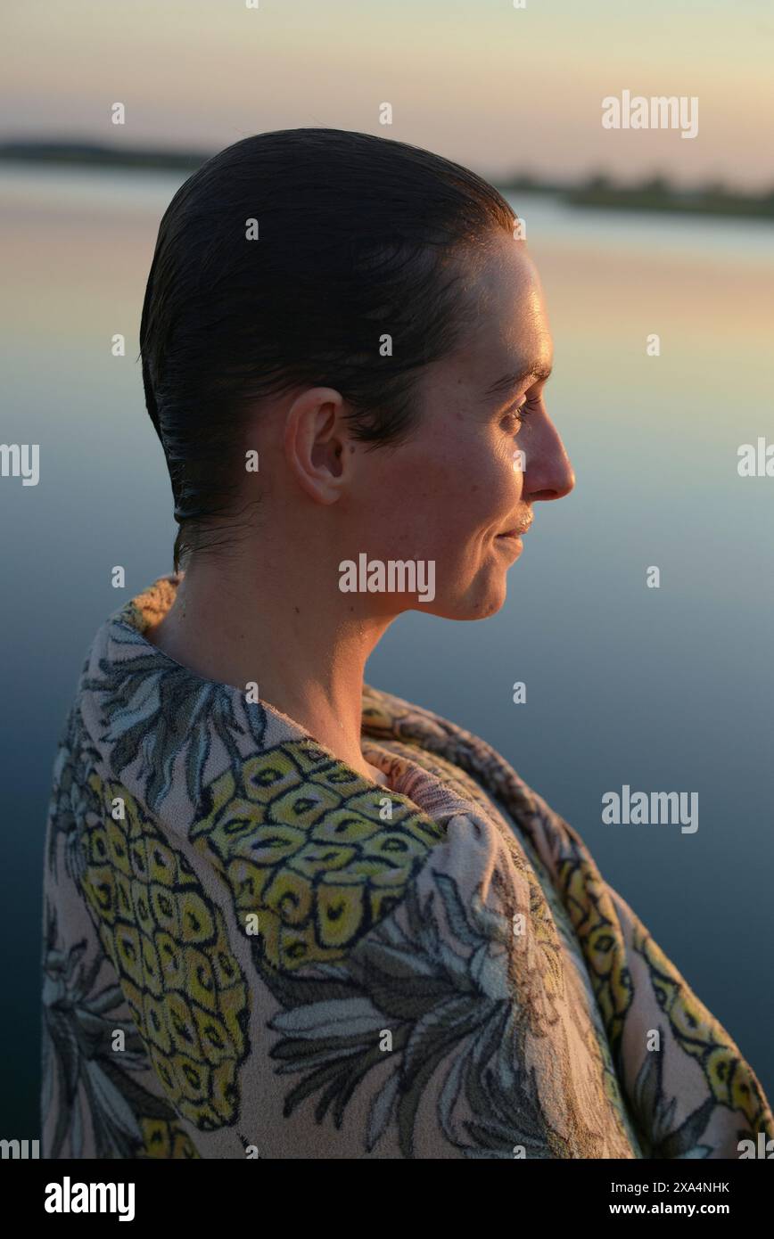 Un profilo laterale di una donna che guarda in lontananza con un corpo d'acqua tranquillo e un tramonto pastello sullo sfondo. Foto Stock