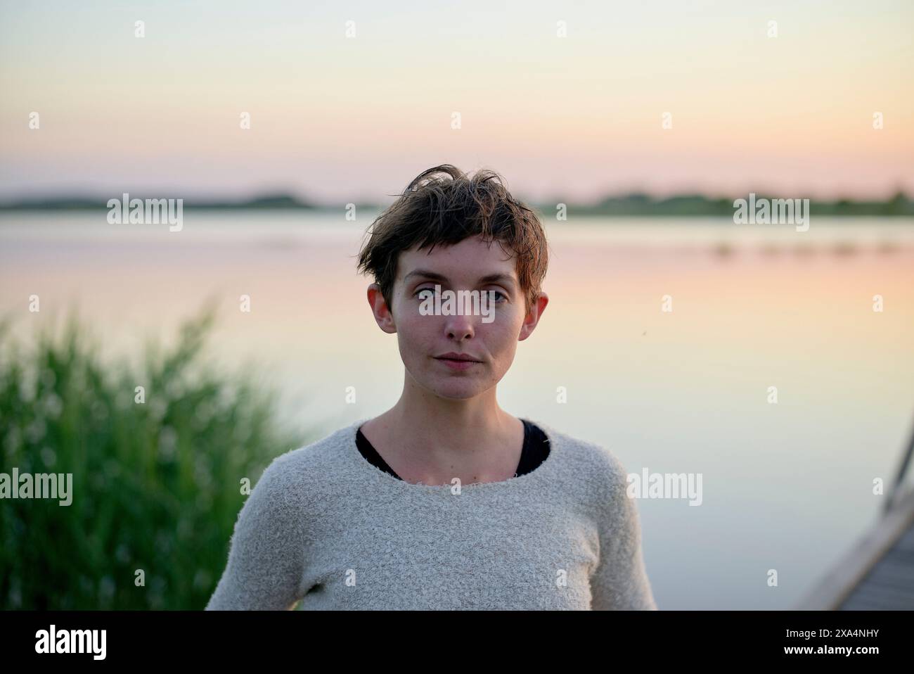 Una giovane donna con i capelli corti si trova accanto a un lago tranquillo durante il tramonto, con un'espressione serena sul viso. Foto Stock