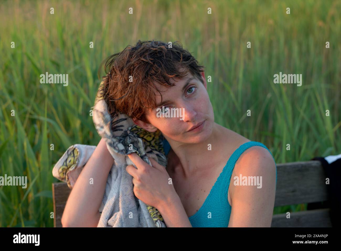 Una persona con i capelli corti e un'espressione contemplativa siede su una panchina, tenendo un asciugamano fantasia, con alte erbe verdi sullo sfondo durante quello che sembra essere il tramonto. Foto Stock