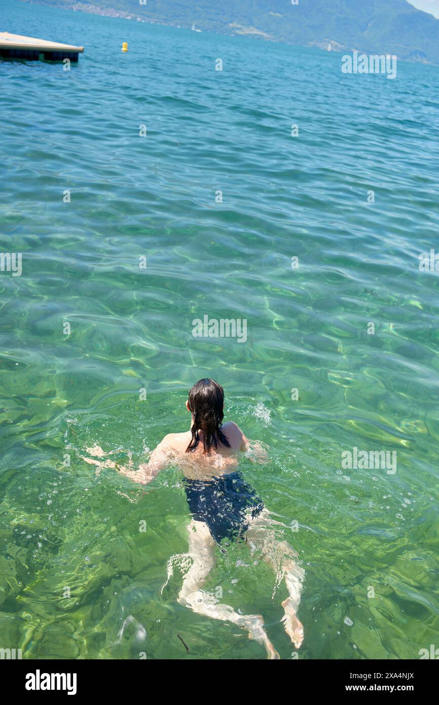 Una persona sta nuotando nelle acque limpide e verdeggianti di un lago con montagne lontane e un cielo soleggiato sopra. Foto Stock