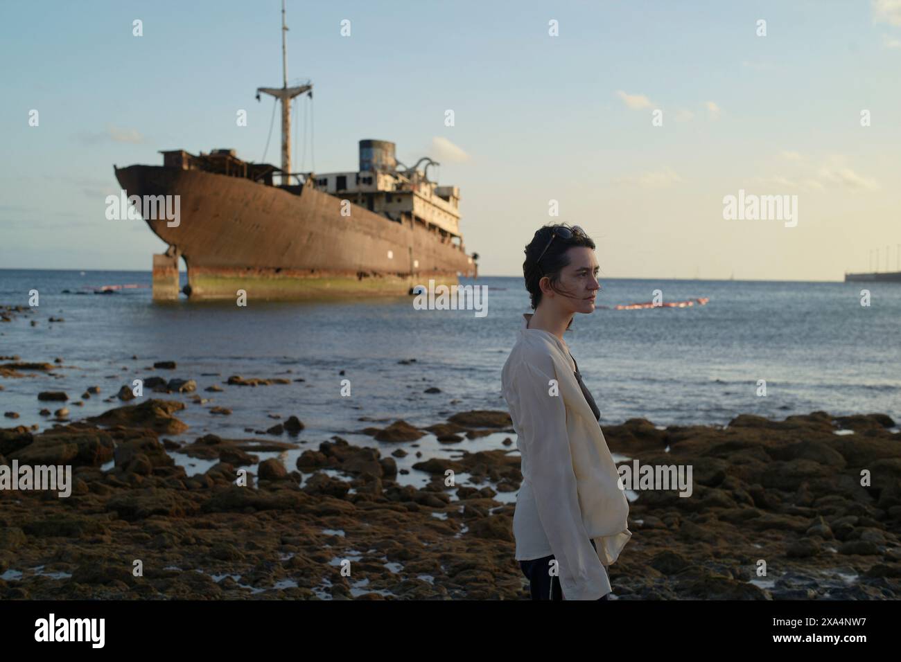Una giovane donna si trova sulla costa rocciosa, di fronte lateralmente con un relitto arrugginito sullo sfondo sotto un cielo nuvoloso. Foto Stock