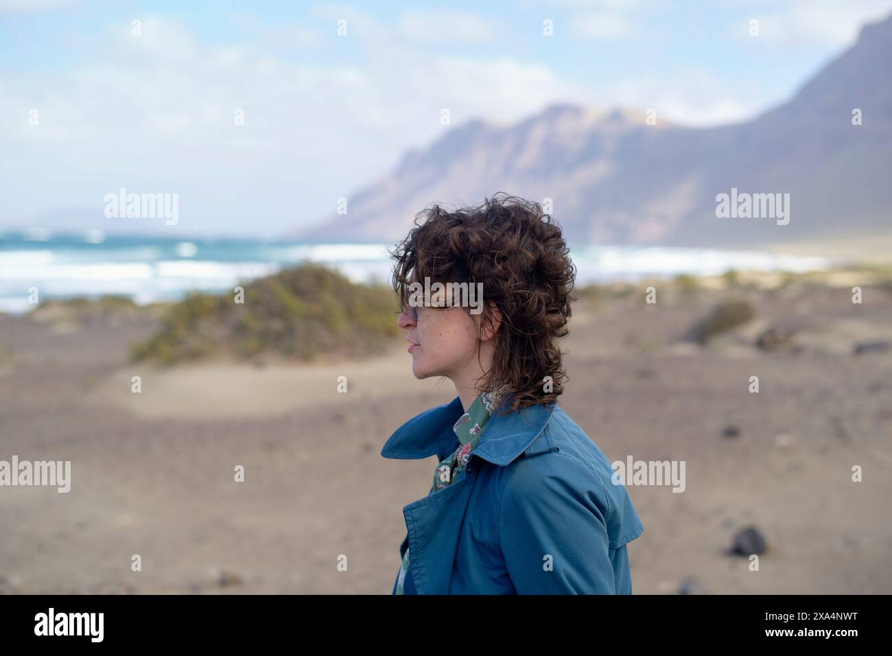 Una giovane donna con i capelli ricci sta in piedi su una spiaggia con montagne sullo sfondo, guardando in lontananza. Foto Stock