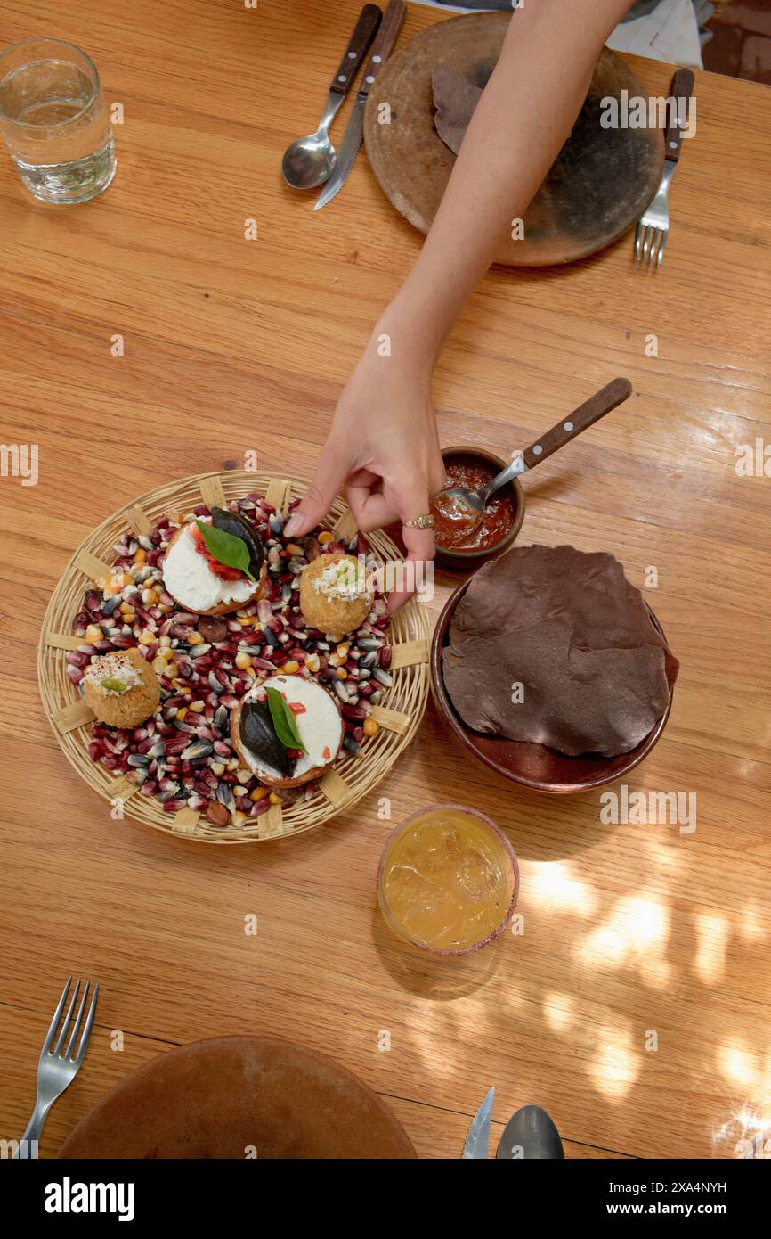 Una persona sta cercando un piatto tradizionale messicano servito su un piatto intrecciato con vari condimenti e salse a lato, su un tavolo di legno. Foto Stock
