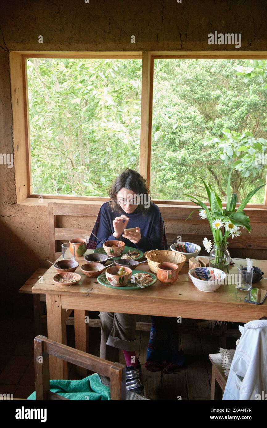 Una giovane donna è seduta ad un tavolo rustico in legno in una sala accogliente con vista sul verde esterno, gustando un pasto da una vasta gamma di ciotole in ceramica. Foto Stock