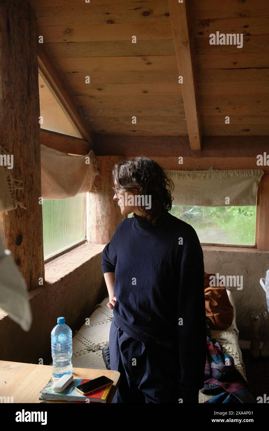 Una giovane donna si trova all'interno di un'accogliente cabina in legno, guardando fuori da una finestra, inondata di luce naturale. Foto Stock