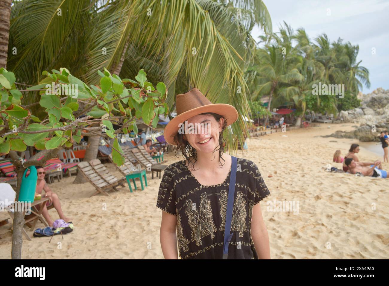 Una persona sorridente che indossa un cappello marrone si erge su una spiaggia sabbiosa con vegetazione e bagni di sole sullo sfondo. Foto Stock