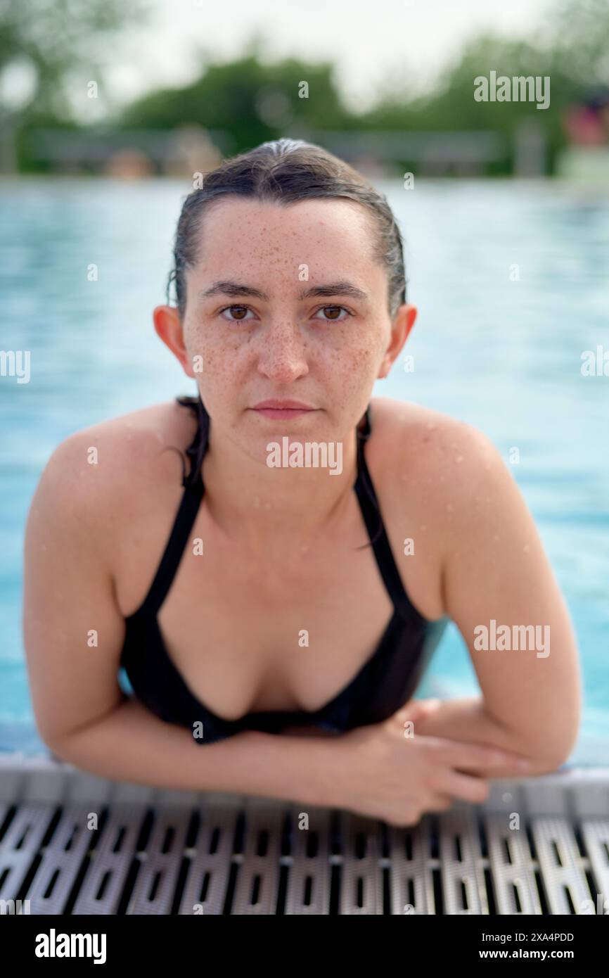 Una donna con i capelli bagnati indossa un costume da bagno nero, che emerge da una piscina con goccioline d'acqua sul viso mentre guarda direttamente nella fotocamera. Foto Stock