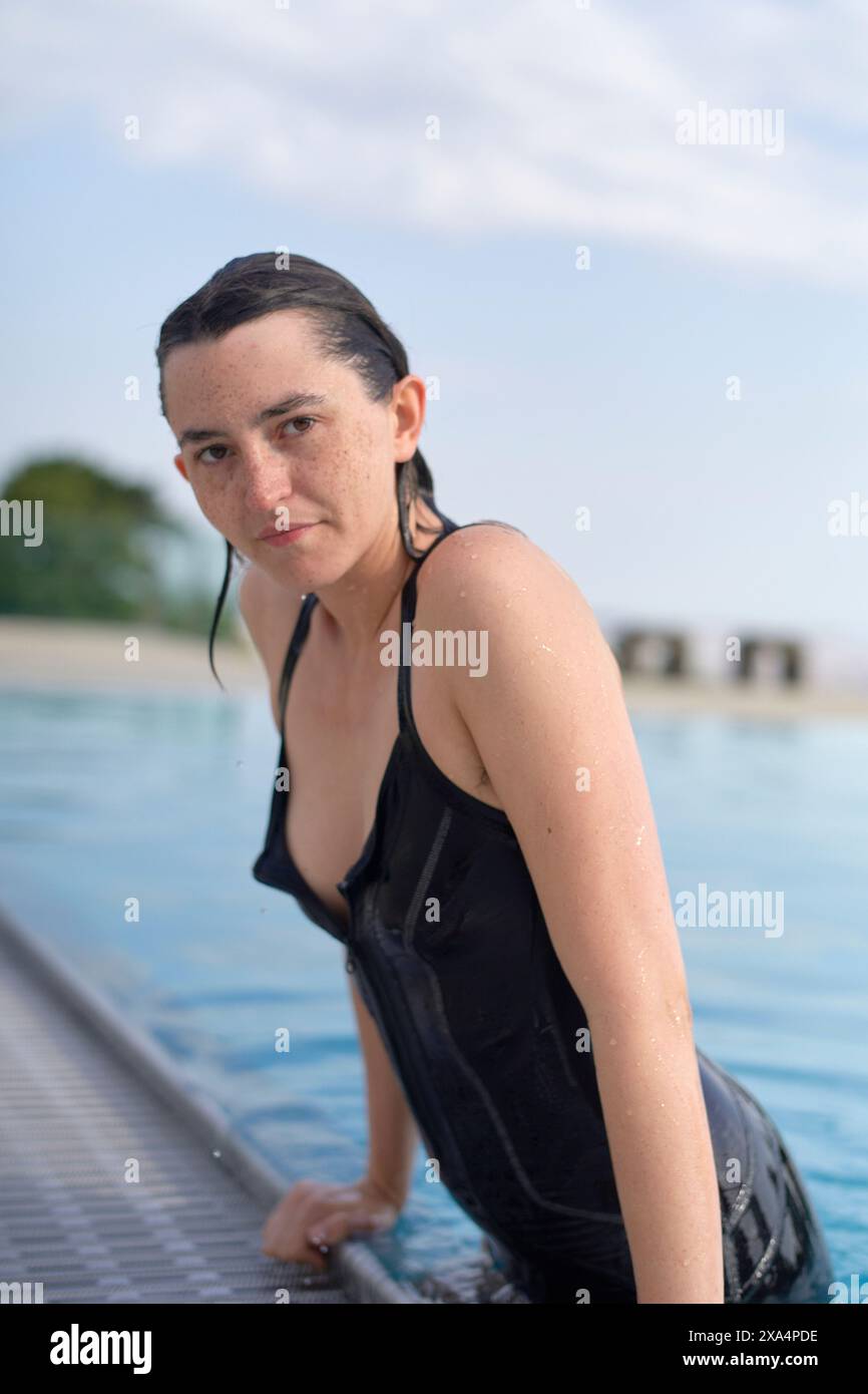 Una donna con un costume da bagno nero bagnato sta emergendo da una piscina, con goccioline d'acqua sulla pelle e un'espressione mirata. Foto Stock