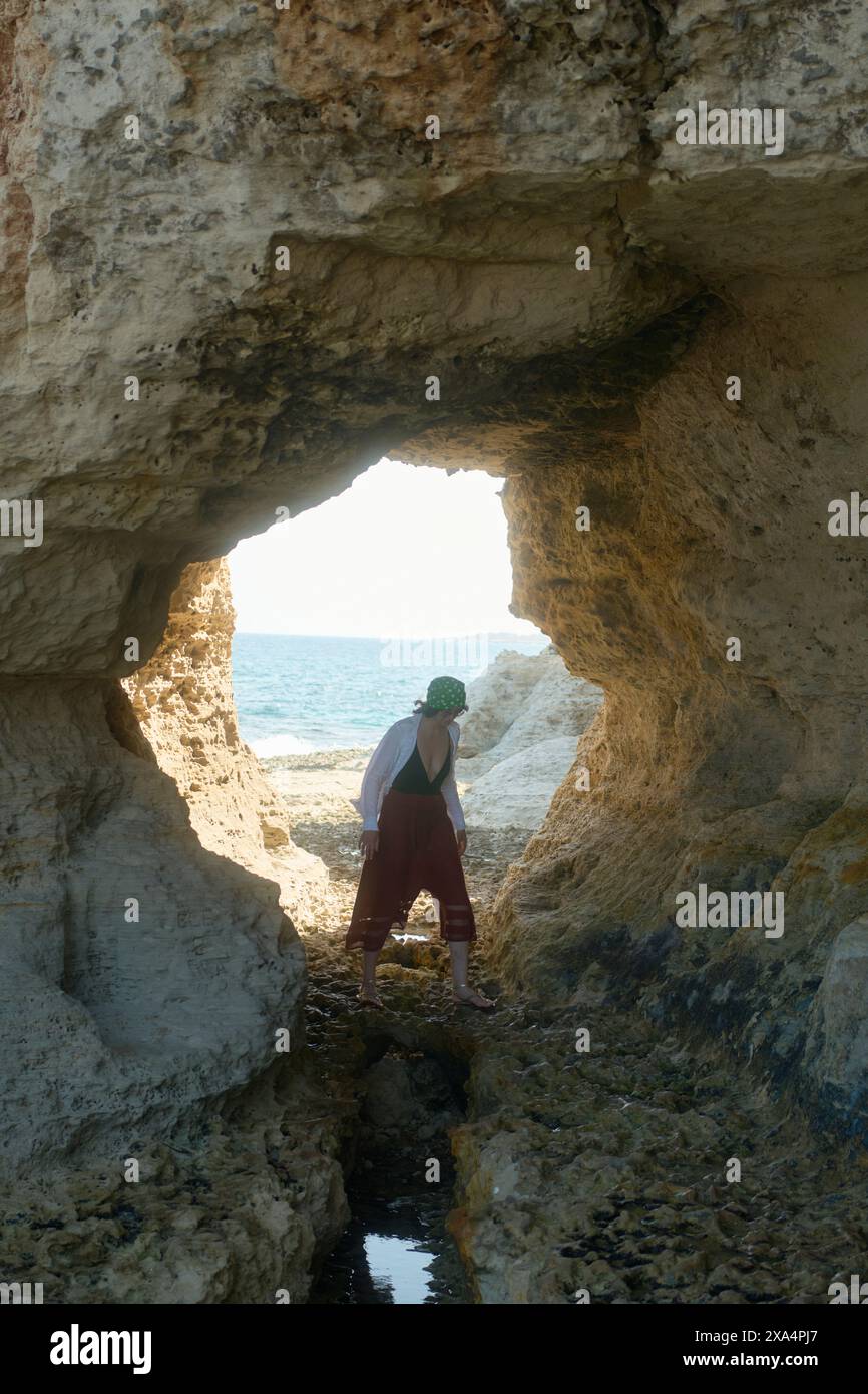 Una persona si trova alla foce di una grotta rocciosa che guarda verso un mare illuminato dal sole. Foto Stock