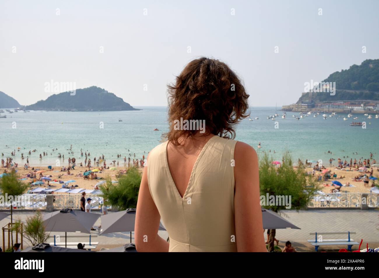 Una donna è in piedi su un balcone, affacciato su una spiaggia affollata con gente che prende il sole e nuota nel mare, con colline in lontananza. Foto Stock