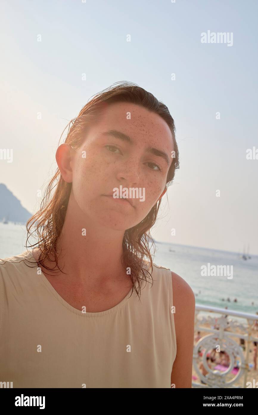 Una giovane donna con lentiggini si erge davanti a una spiaggia soleggiata, i capelli si sono spostati da un lato, con la luce del sole che proietta un bagliore morbido sul viso. Foto Stock