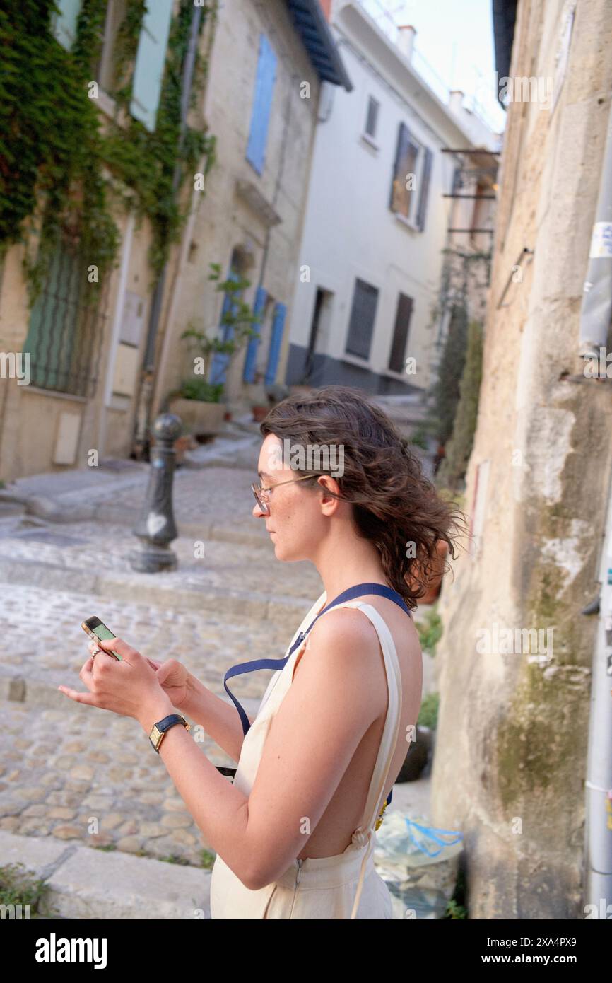 Una donna sta in piedi su una strada acciottolata in una città vecchia, tenendo in mano uno smartphone e sorridente, con edifici e vegetazione sullo sfondo. Foto Stock