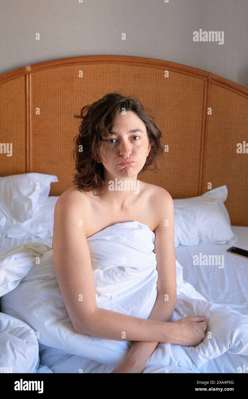 Una persona è seduta in un letto con lenzuola bianche, avvolta da una coperta, e guarda direttamente la telecamera con un'espressione neutra. Foto Stock