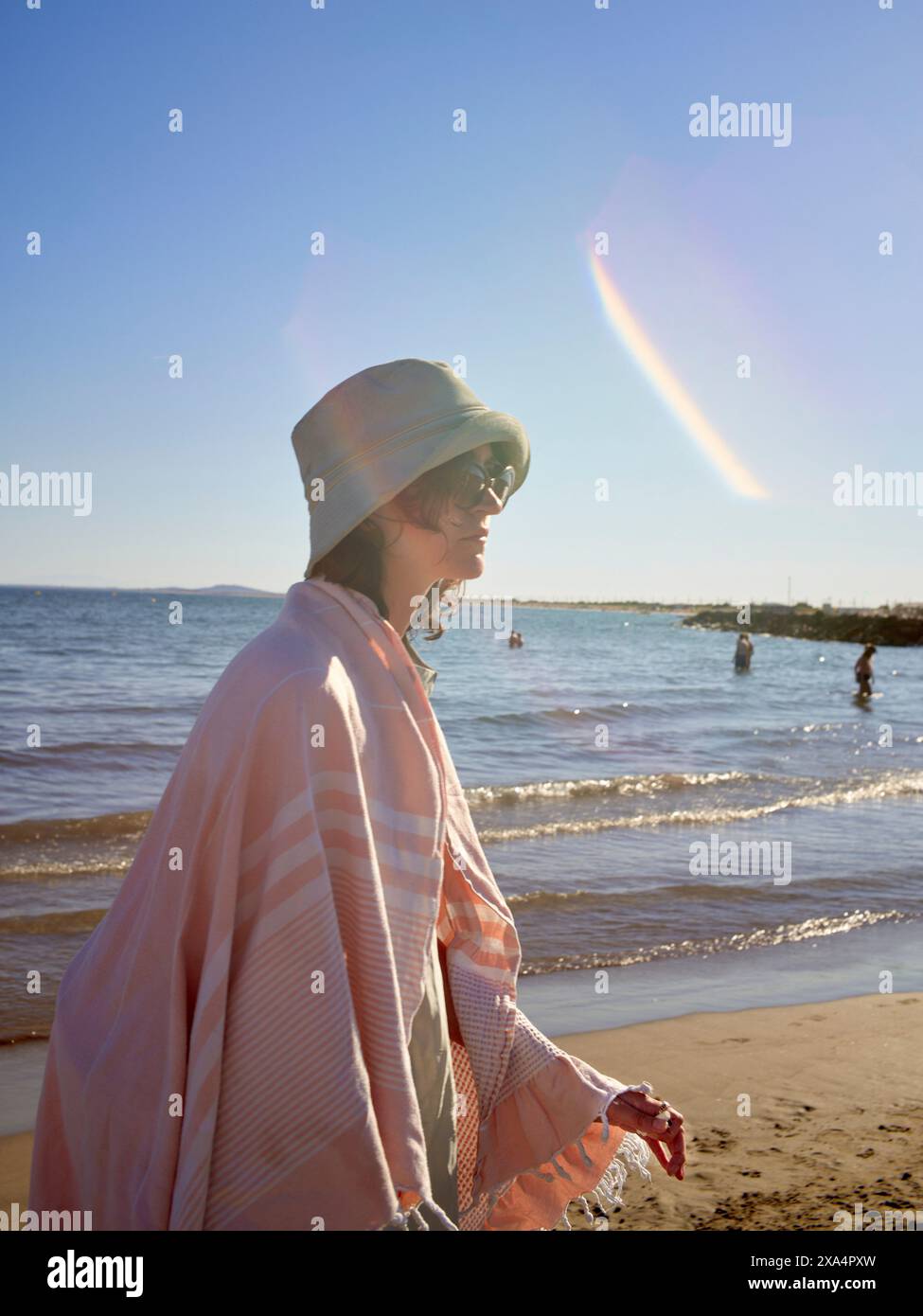Una giovane donna sta sulla spiaggia drappeggiata da una coperta a righe rosa, indossa un cappello da secchio, con il sole che proietta una calda luce e un bagliore di lenti. Foto Stock