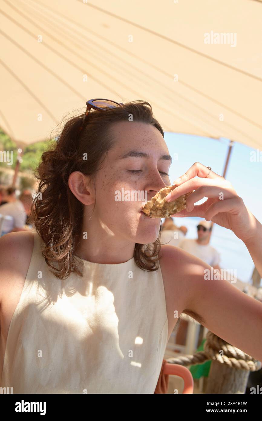 Una persona sta gustando un pasto all'aperto, prendendo un boccone di cibo sotto un'area ombreggiata con un cielo limpido e soleggiato sullo sfondo. Foto Stock