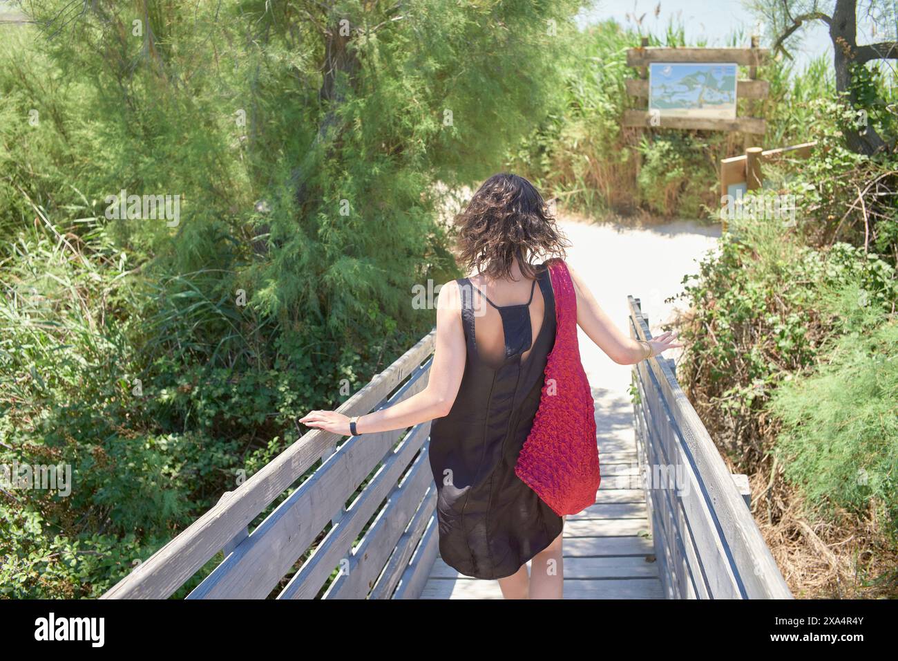 Una donna con un abito nero e una borsa rossa cammina lungo una passerella di legno verso un sentiero sabbioso fiancheggiato da arbusti verdi sotto un cielo luminoso. Foto Stock