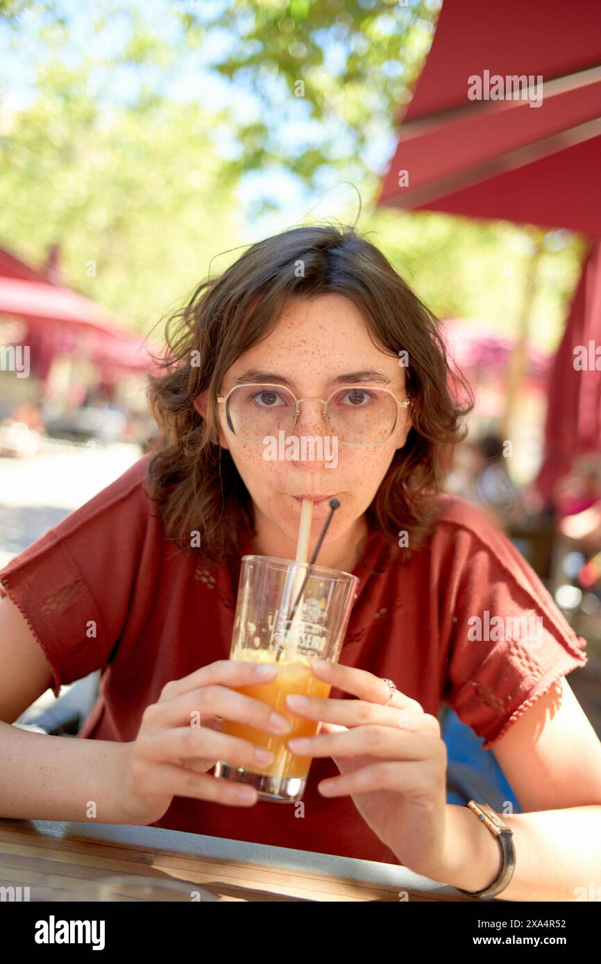 Una giovane donna è seduta ad un tavolo da caffè all'aperto, tenendo un bicchiere di succo d'arancia e guardando direttamente la macchina fotografica. Foto Stock