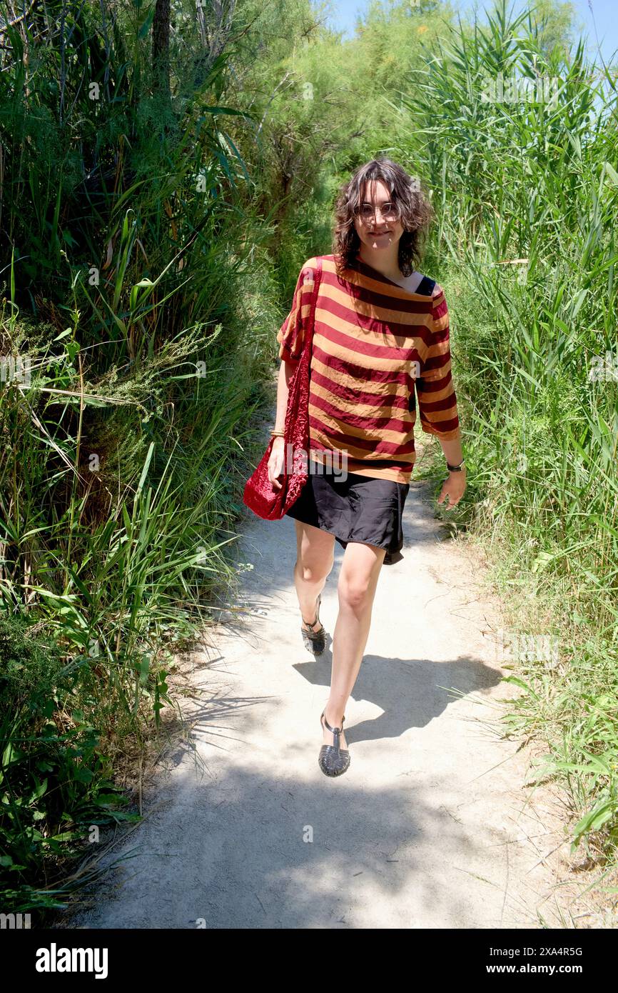 Una persona sta camminando lungo un sentiero circondato dal verde, portando con sé una borsa rossa e indossando un maglione a righe e pantaloncini corti. Foto Stock