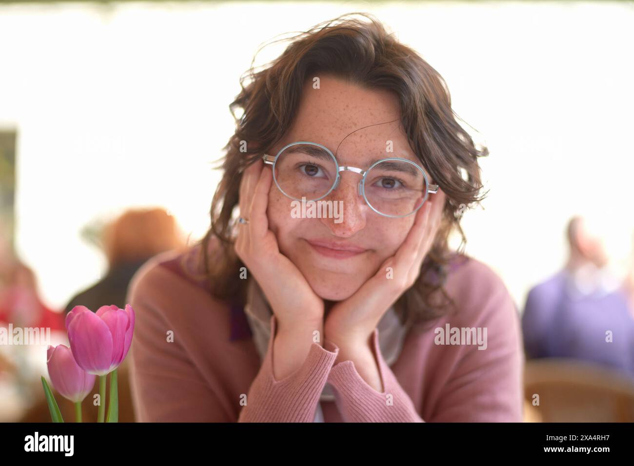 Donna sorridente con occhiali rotondi che appoggiano il mento sulle mani accanto a un tulipano rosa Foto Stock
